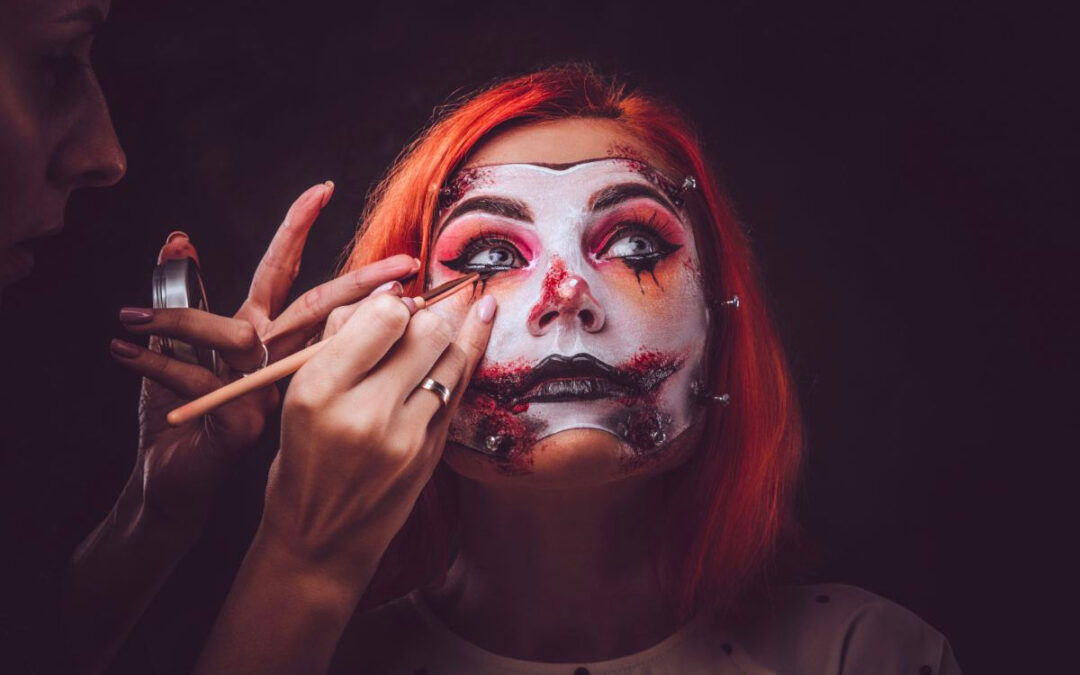 Une jeune femme rousse se fait maquiller pour Halloween. Les mains de la maquilleuse dessinent du faux sang, des larmes noires et une bouche noire ensanglantée avec des cicatrices, rappelant le style du Joker dans Batman.